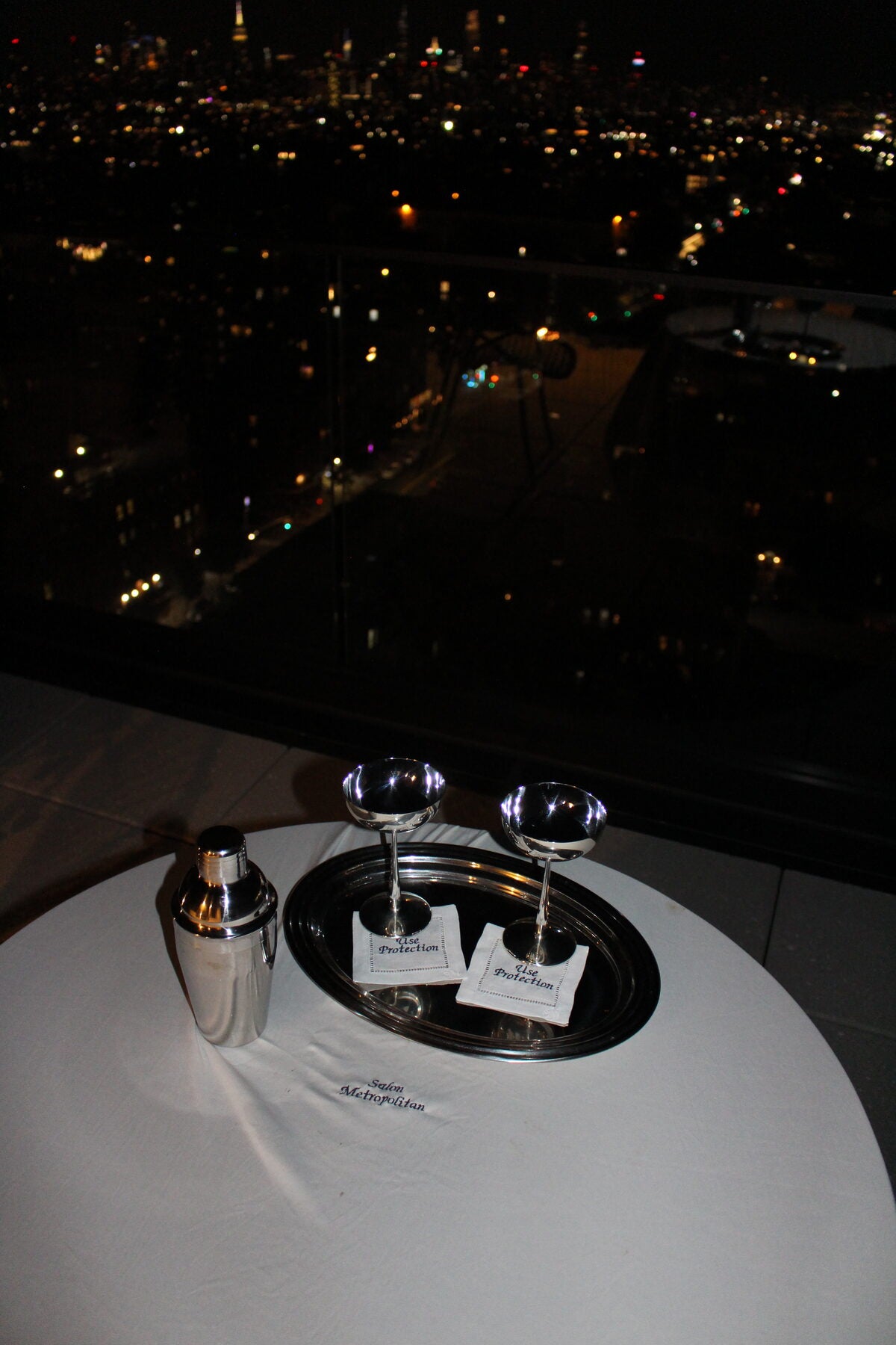 Cocktail shaker and coupe glasses with Use Protection napkins, overlooking the city at night
